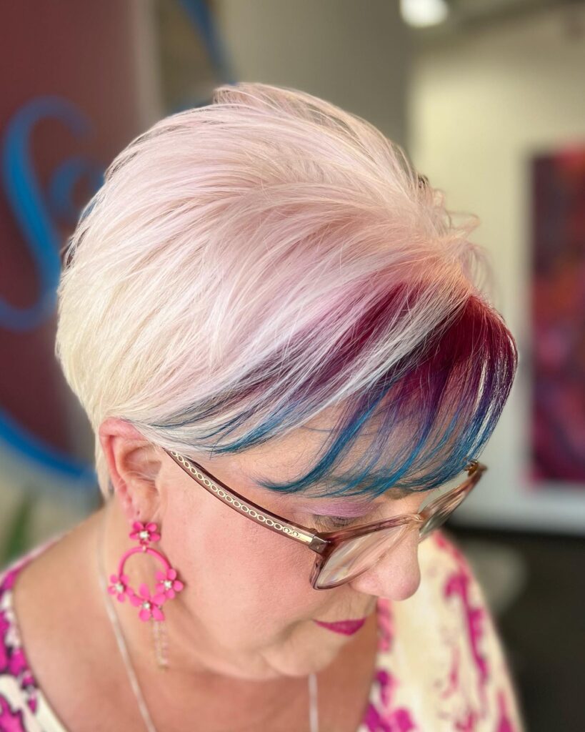 A woman over 60 with a platinum and pastel pink pixie cut featuring vibrant magenta and turquoise highlights in her side-swept fringe.