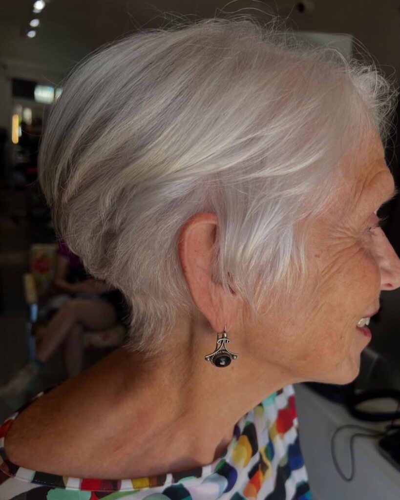 A woman over 60 with a wispy silver-white pixie-bob cut featuring long tucked layers and a feathered neckline.