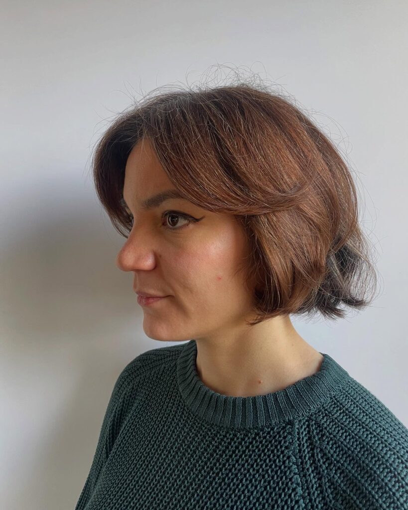 A woman in side profile showcasing a jaw length choppy cinnamon brown bob hairstyle with sweeping winged layers and a gentle lift.