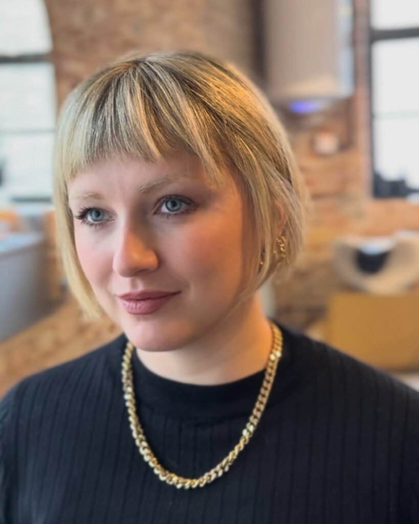 A woman looking to the side wearing a jaw length warm blonde choppy bob with a short wispy fringe.