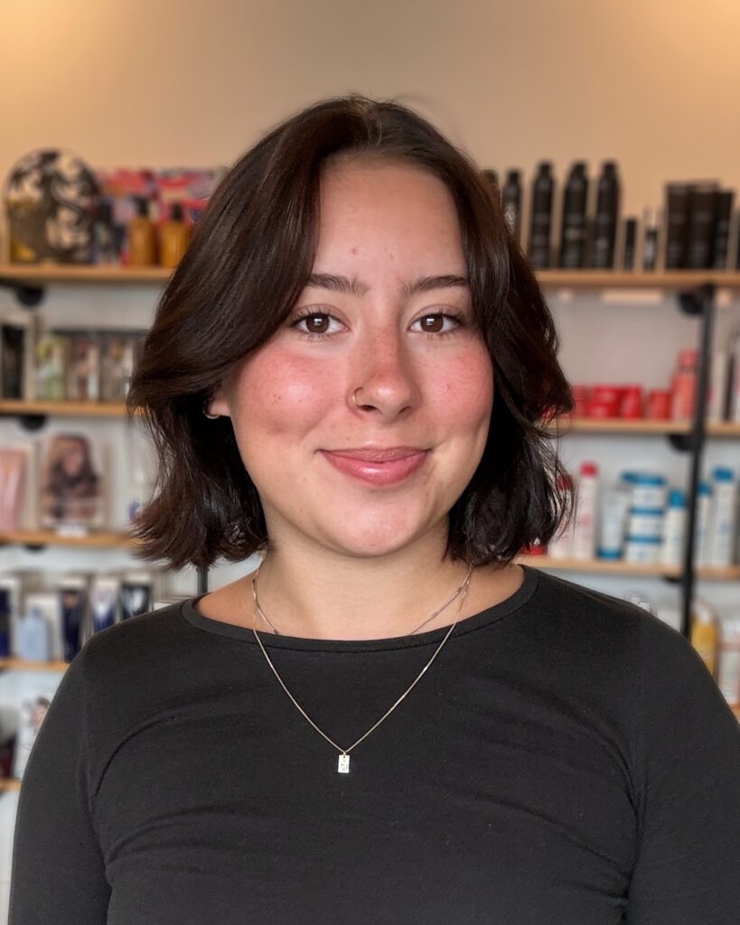 A smiling woman showcasing a jaw length choppy dark brown bob hairstyle with layered bangs that sweep outward to the sides.