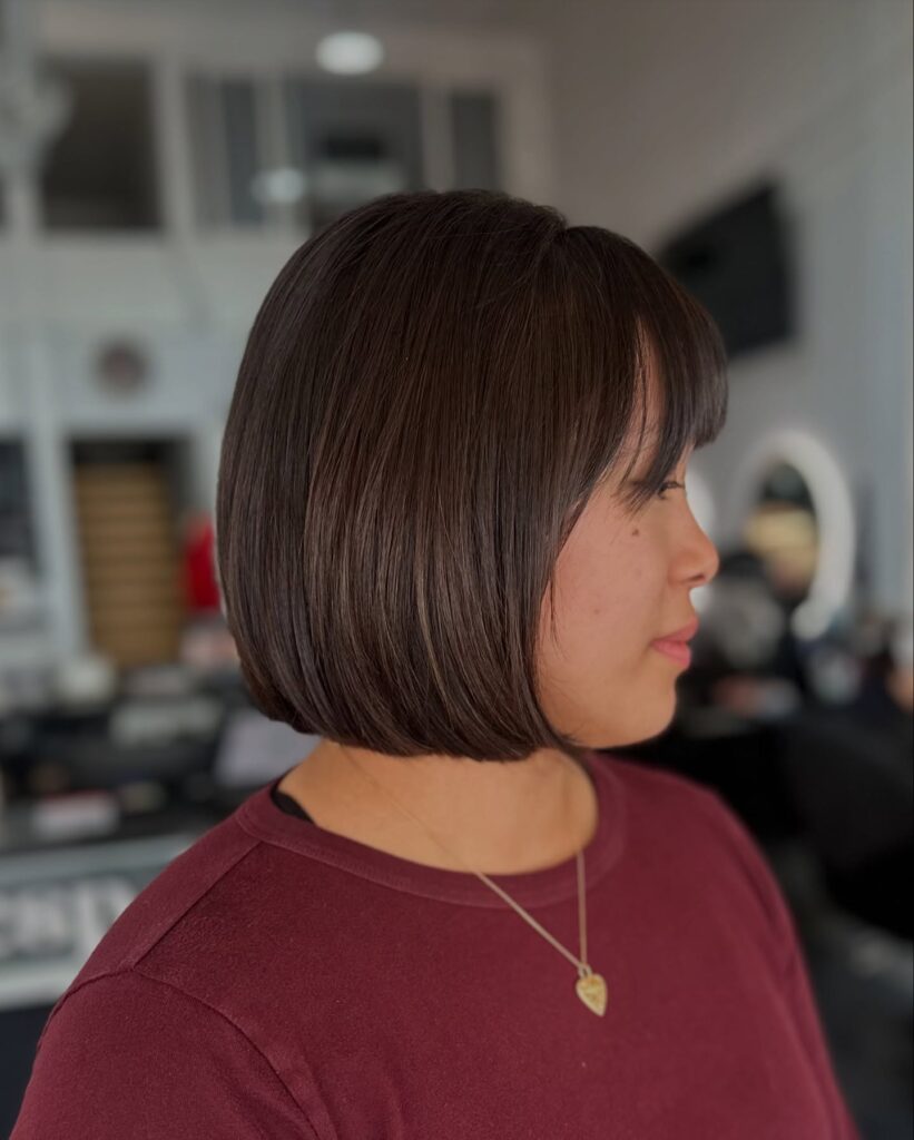 A woman in side profile showcasing a jaw length choppy mahogany brown bob hairstyle with a wispy fringe and a rounded under-beveled shape.