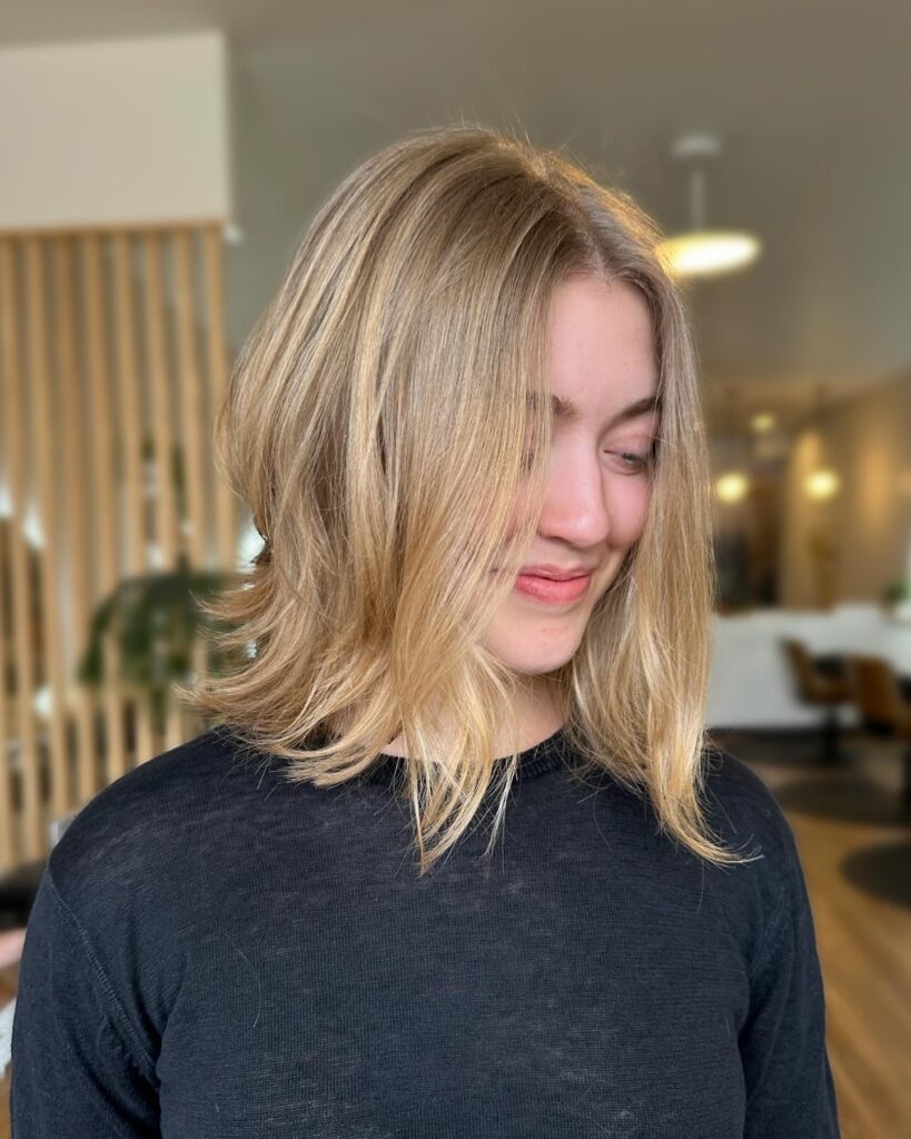 A smiling woman looking down and to the side showcasing a jaw length choppy honey blonde bob hairstyle with flicked layers and a gentle lift.