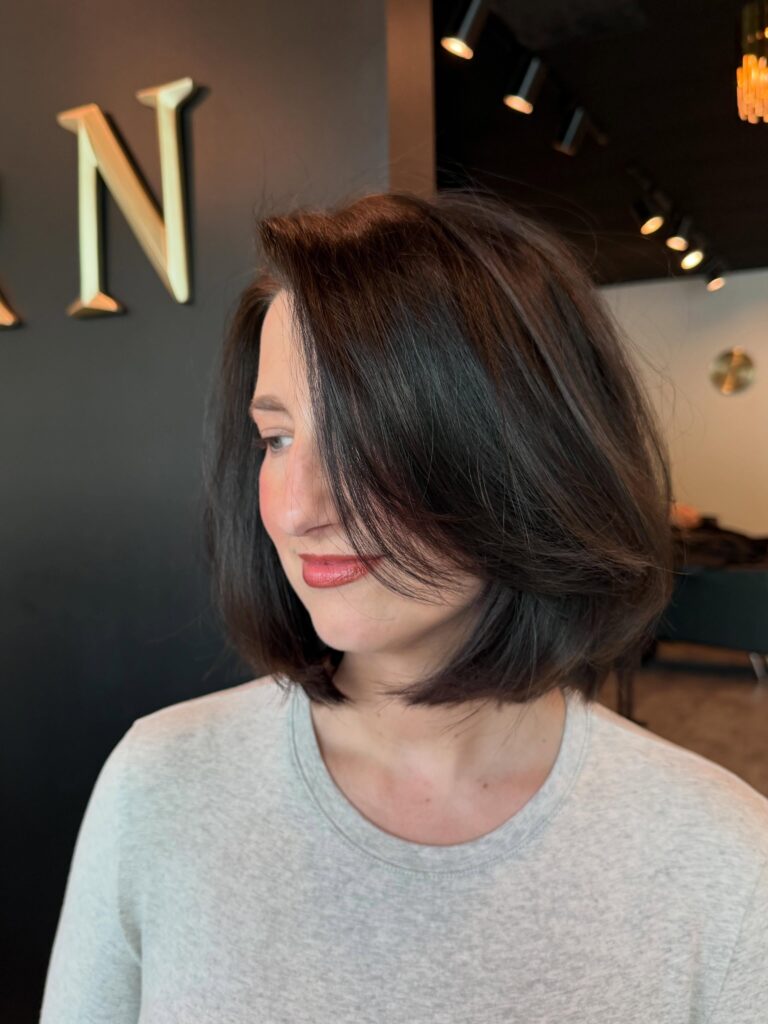A woman looking down to the side, showcasing a voluminous dark brown collarbone-length long bob haircut with a deep side part and soft curved layers.
