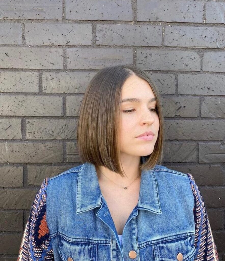 A woman with a straight, neck-length chestnut brown bob haircut featuring a center part and sharp blunt edges.