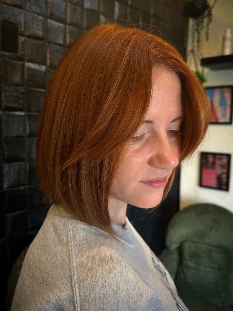 A close-up side profile view of a woman showcasing a straight, jaw-length chocolate brown angled bob haircut with ultra-clean perimeter lines.