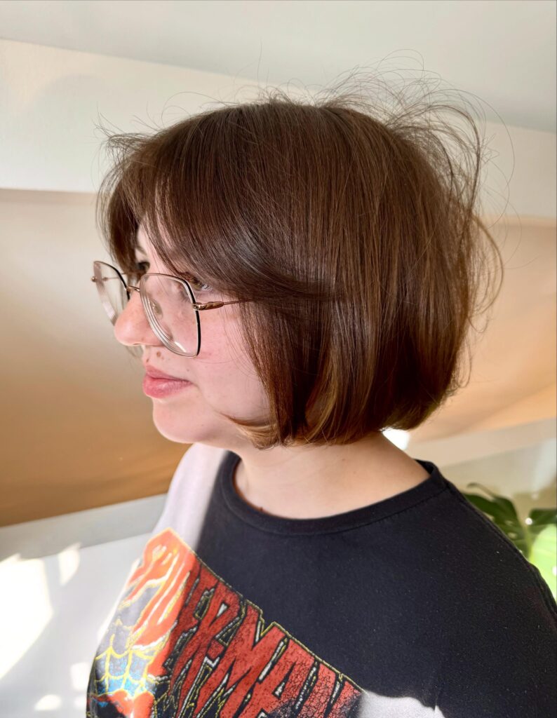 A side profile view of a woman showcasing a straight honey blonde neck-length bob haircut with golden highlights and a gentle forward angle.
