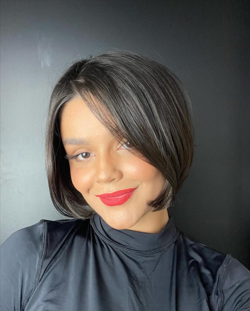 A smiling woman with a sleek, jaw-length jet black angled bob haircut featuring a deep side part and long side-swept bangs.