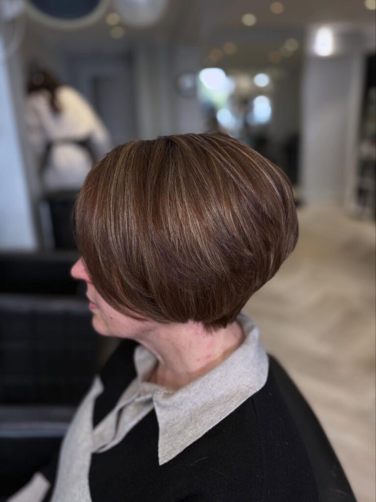 A side profile view of a woman showcasing a straight, stacked chestnut brown bob haircut with fine caramel highlights and a volumetric back.