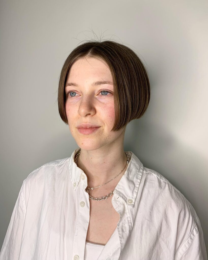 A young woman with blue eyes showcasing a sleek, center-parted ash brown micro bob haircut that angles sharply upward toward the back.