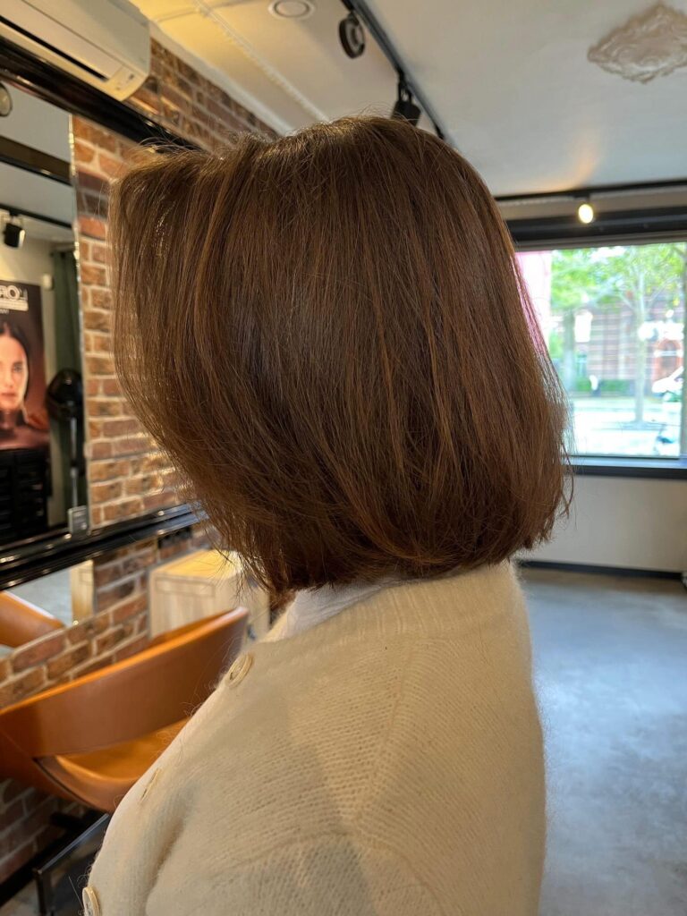 A side profile view of a woman showcasing a straight, shoulder-length caramel brown long bob haircut with a subtle forward angle.