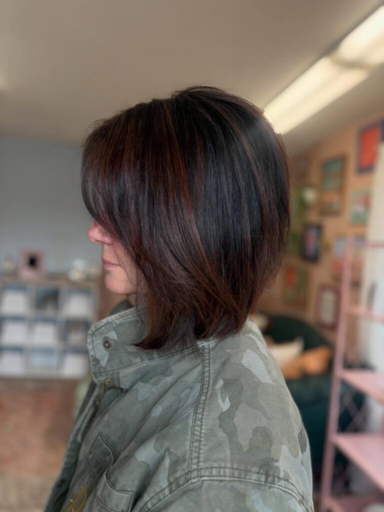 A side profile view of a woman showcasing a sleek, straight mahogany brown bob haircut with a sharp forward angle.
