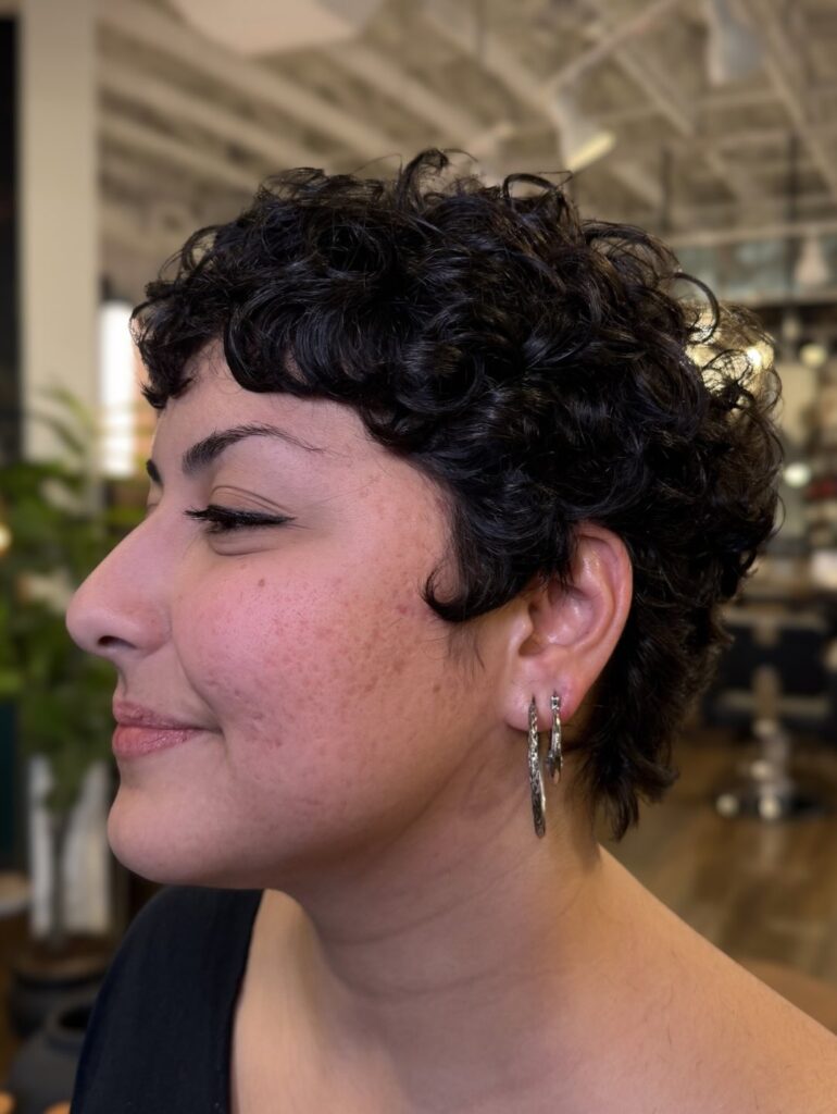 Short dark curly pixie haircut with soft baby bangs and layered texture around the crown.