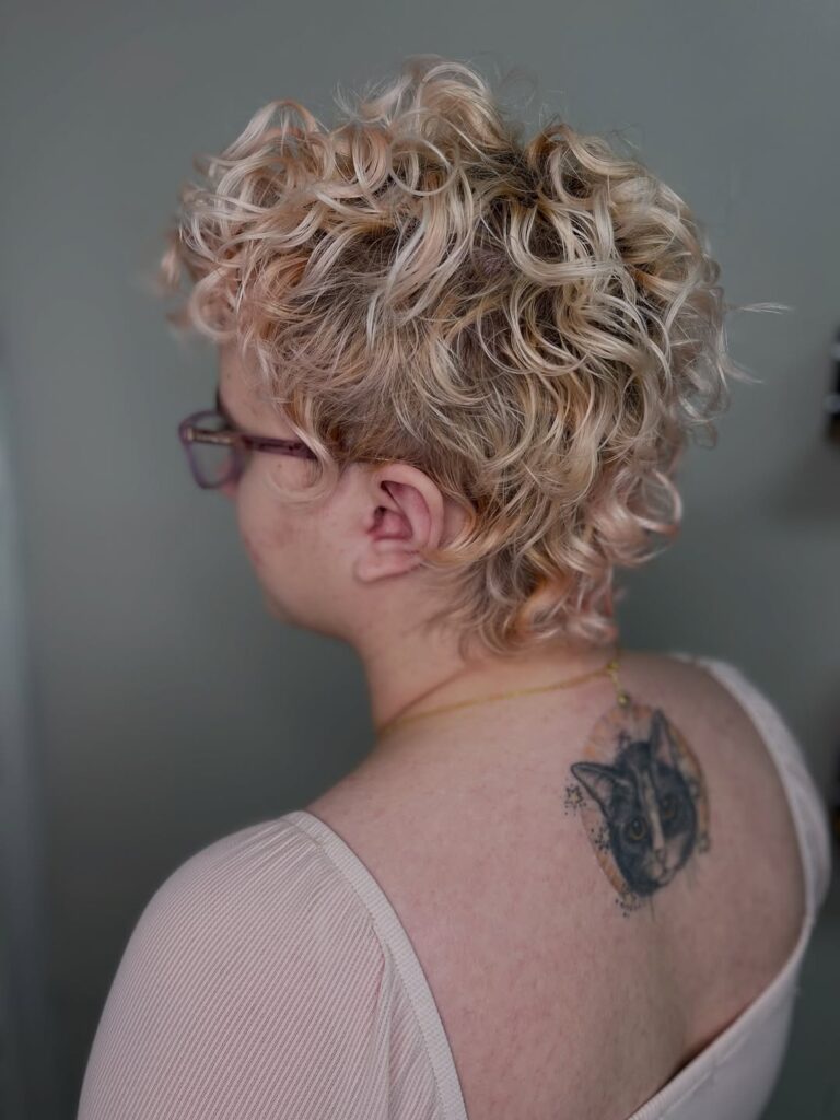 Blonde curly pixie haircut with layered curls and a softly tapered back.