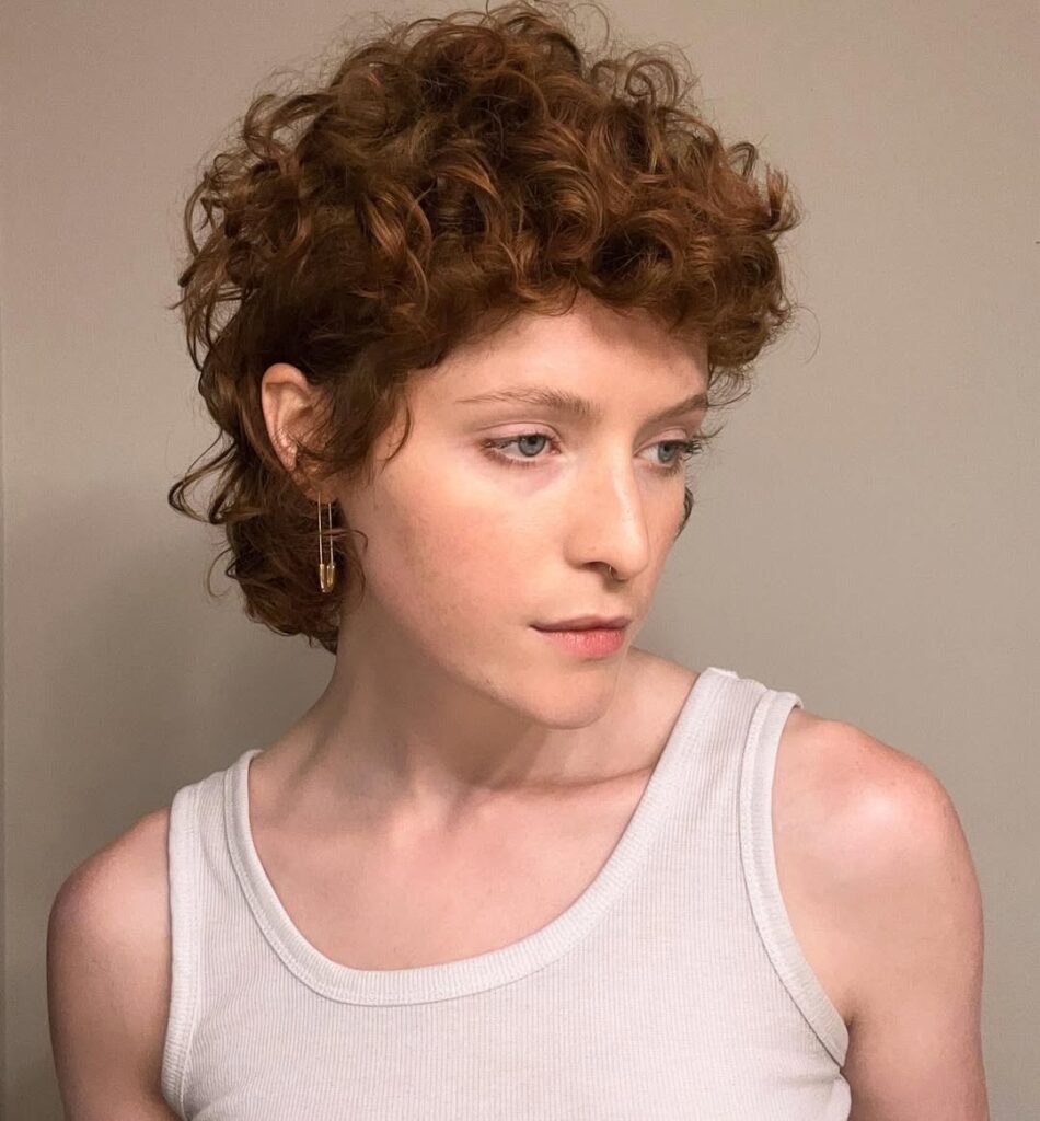 Short copper curly pixie hairstyle with soft layers, fluffy volume on top, and curled pieces around the nape.