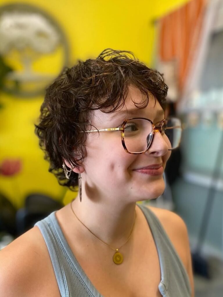 Short dark curly pixie hairstyle with wispy bangs and soft curls styled around glasses.