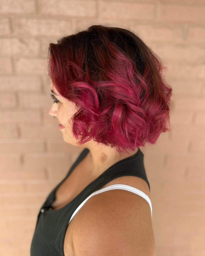 A raspberry red curled bob with dark roots and choppy layered texture.