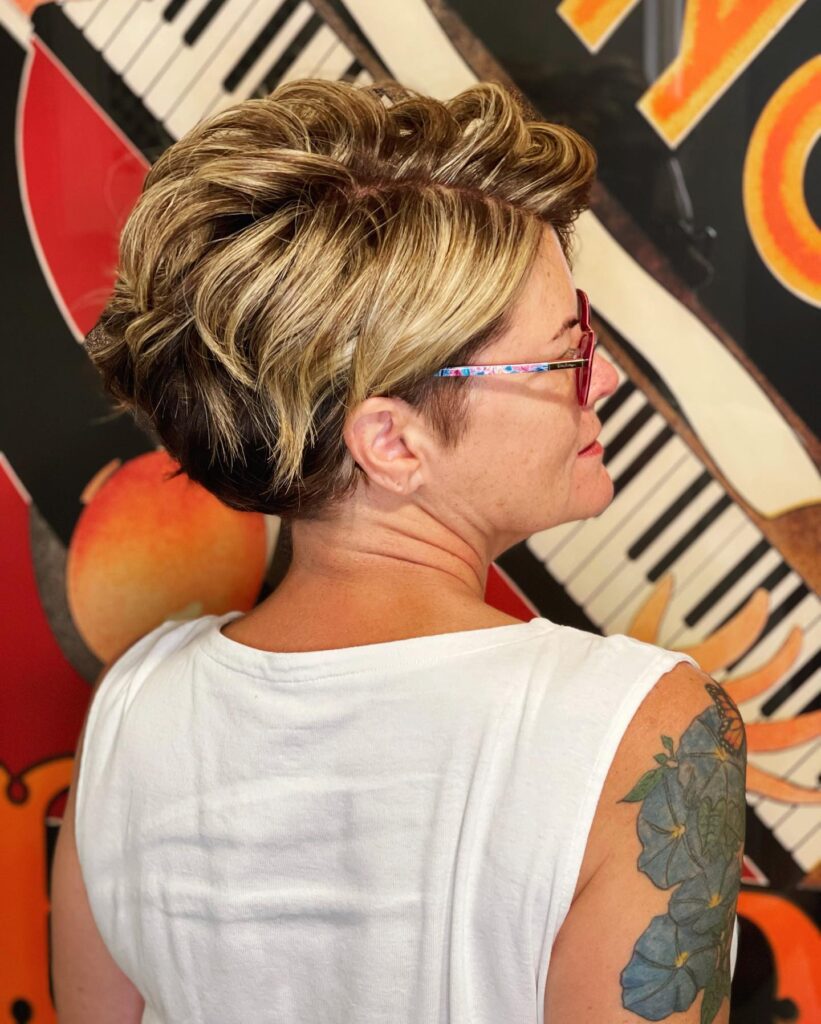 A woman with thick multi-tonal blonde hair wears a wavy pixie cut with significant volume on top and a tapered back.