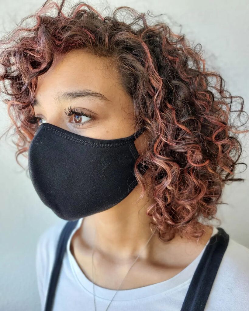 Layered curly bob with soft coils and muted rose pink highlights throughout.