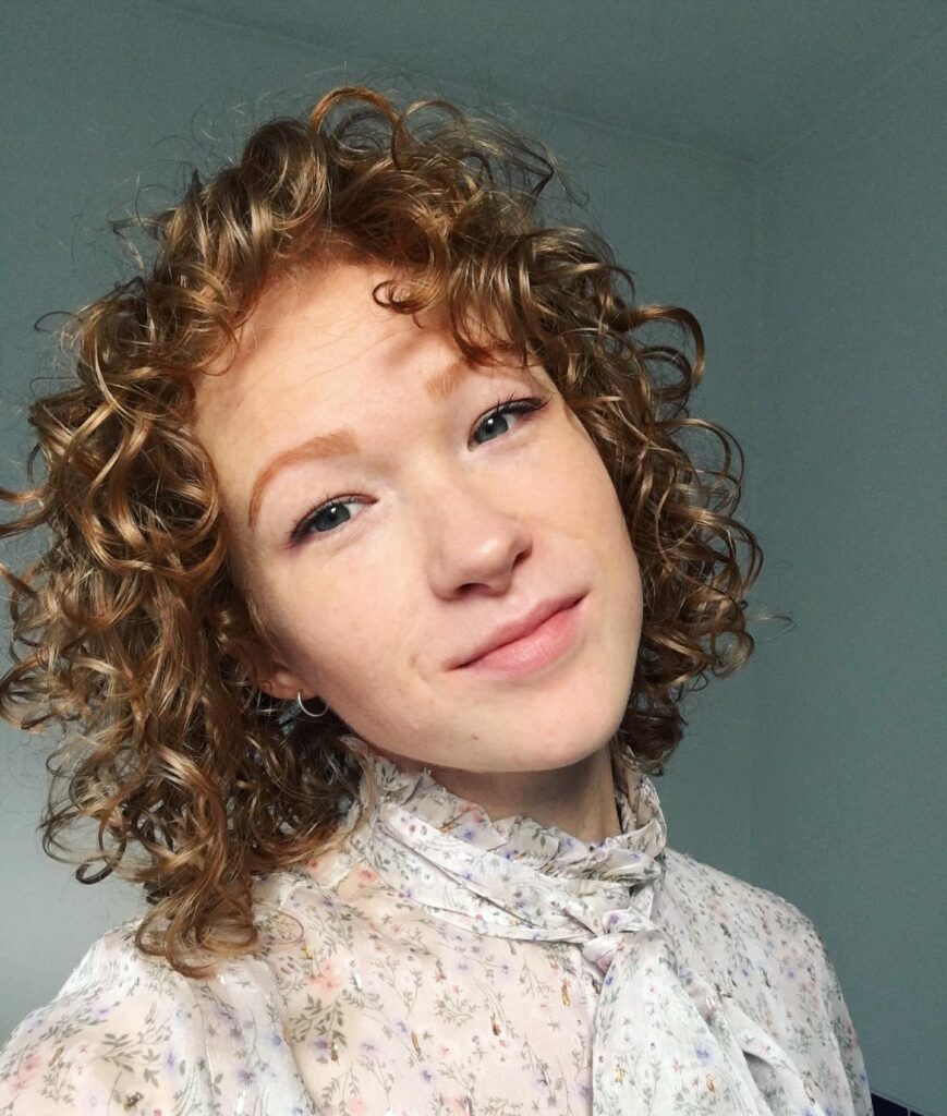 Short layered bob with tight spiral curls, soft curly bangs, and warm caramel brown tones.