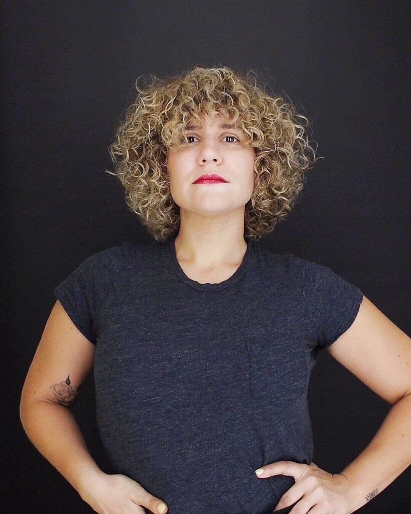 Golden blonde curly bob with rounded layers and full curly bangs.