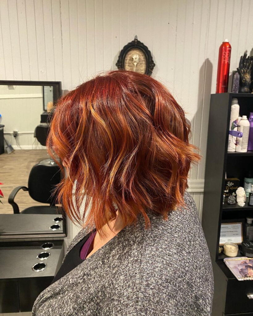A woman with a shoulder-length layered bob styled in loose waves with a mix of deep red and copper highlights.