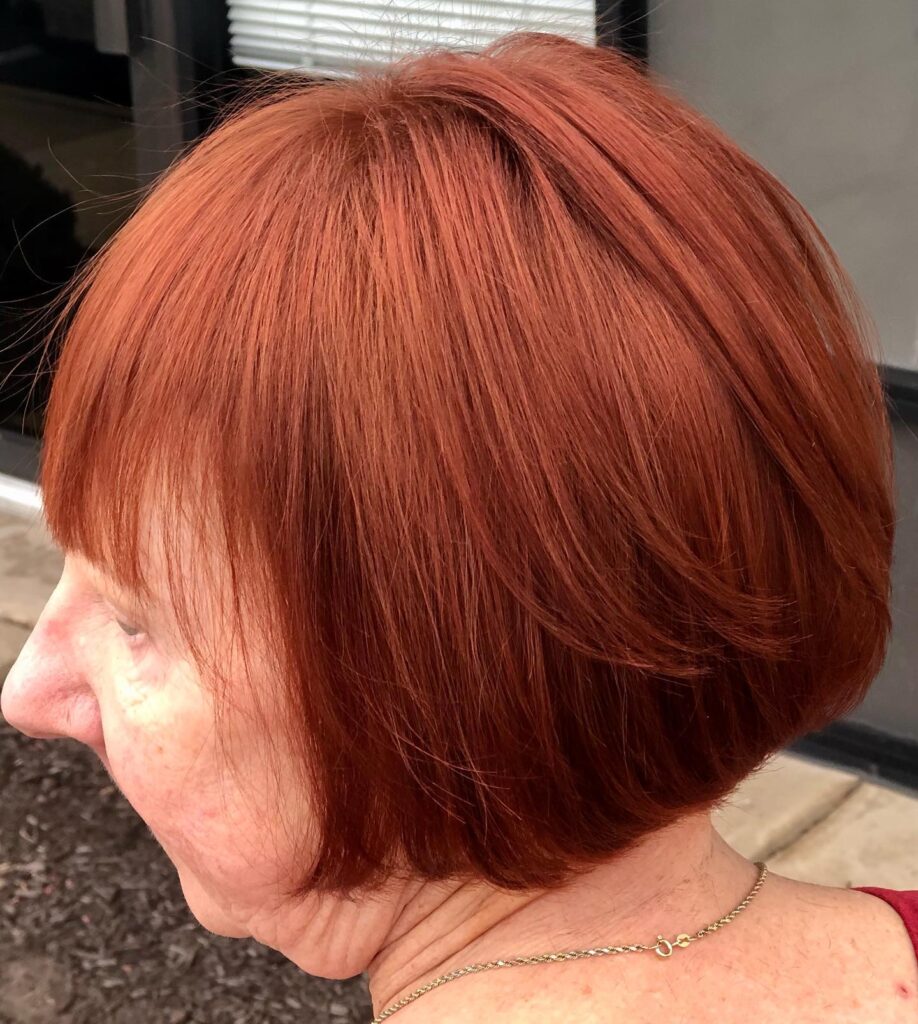 A woman with a sleek jaw-length bob haircut and wispy bangs in a vibrant, solid red-copper color.