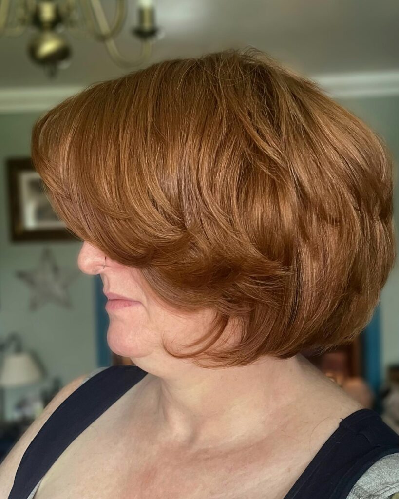 A woman in profile wearing a voluminous chin-length bob in a warm terracotta copper shade with feathered layers.