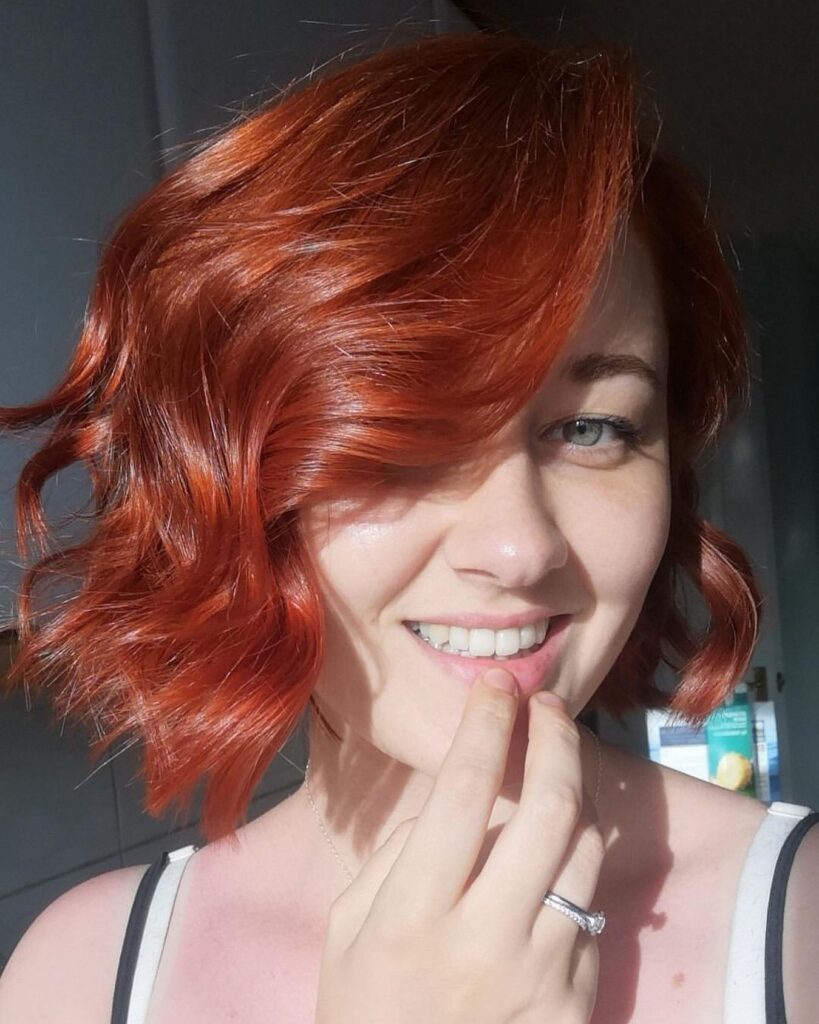 A smiling woman with a wavy, neck-length bob in a glossy crimson copper color.