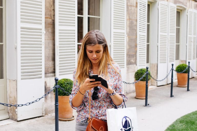 The Best Texts Are Usually the Ones That Feel a Little Unscripted A woman with long, wavy hair checks her phone while standing outside a building with white shutters, holding a shopping bag and wearing a floral blouse.