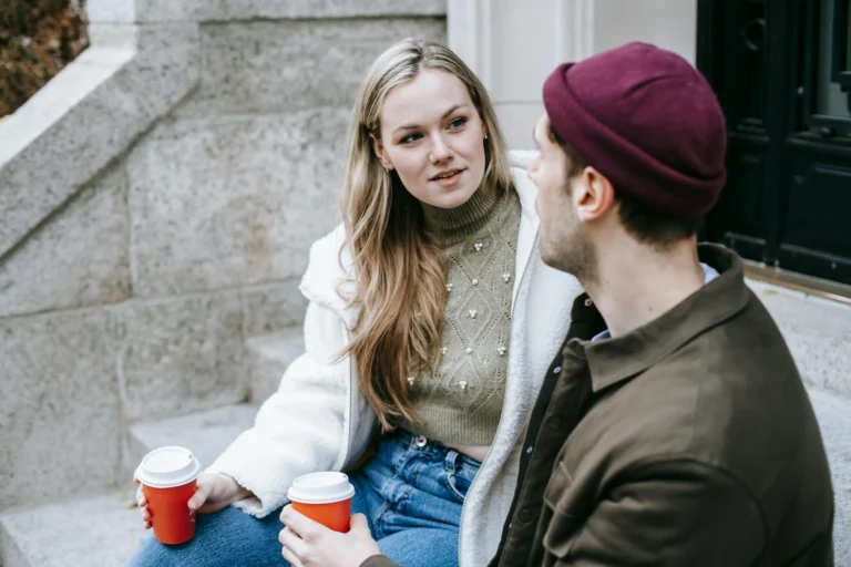 Good Conversation Gets Better When the Questions Feel Less Performative A young woman with long, wavy hair wears a cozy sweater and a white jacket, engaged in conversation with a man in a beanie, both holding coffee cups.