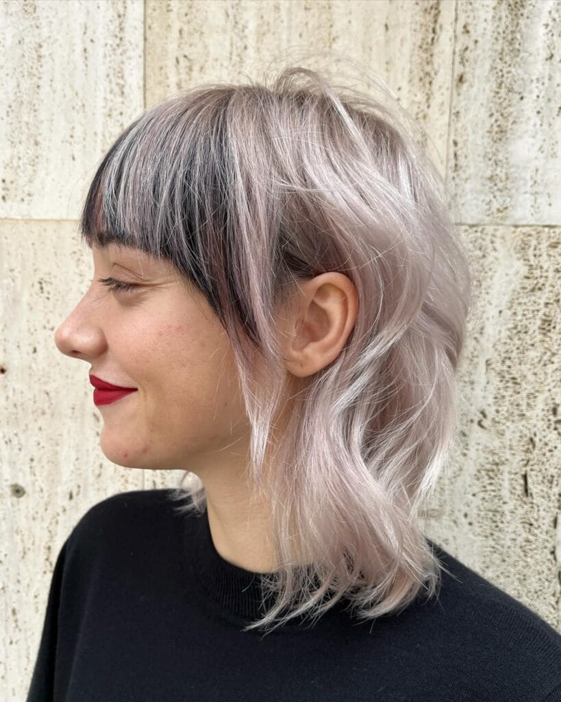 A woman with an icy lavender layered mullet featuring contrasting dark, blunt bangs and wavy shoulder-length ends.