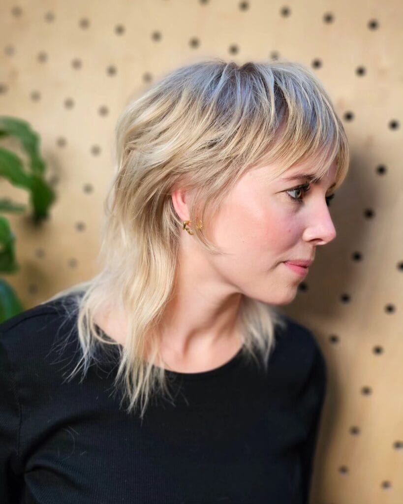 A woman with a soft sandy blonde mullet featuring shaggy feathered layers and a light fringe.