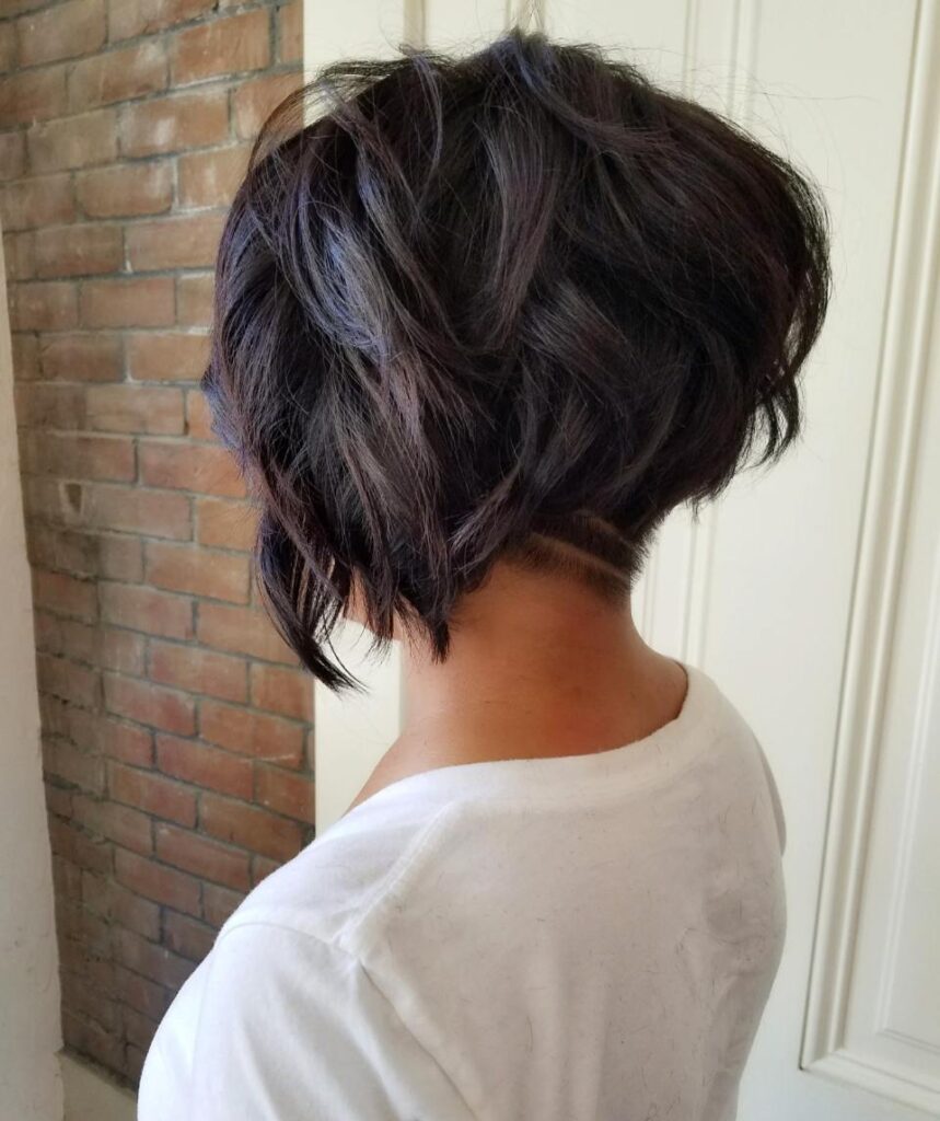 Rear view of a woman with a choppy dark brown inverted wavy bob haircut featuring a stacked back and a shaved undercut with two horizontal lines.