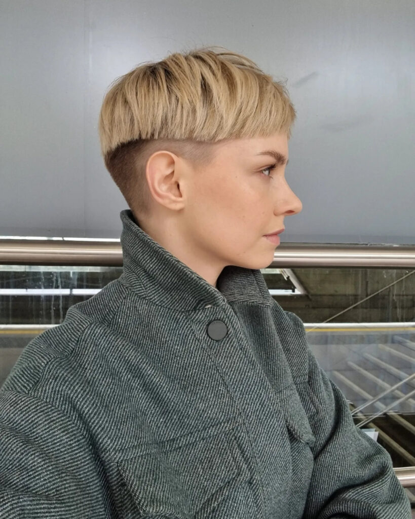 A side profile of a woman with a sandy blonde bowl cut featuring a blunt perimeter and a high shaved skin-fade undercut.