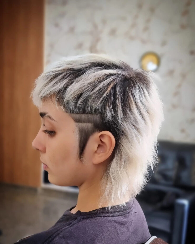 A side profile of a woman with a choppy white-blonde mullet featuring dark roots and a shaved-temple undercut with a single horizontal line.