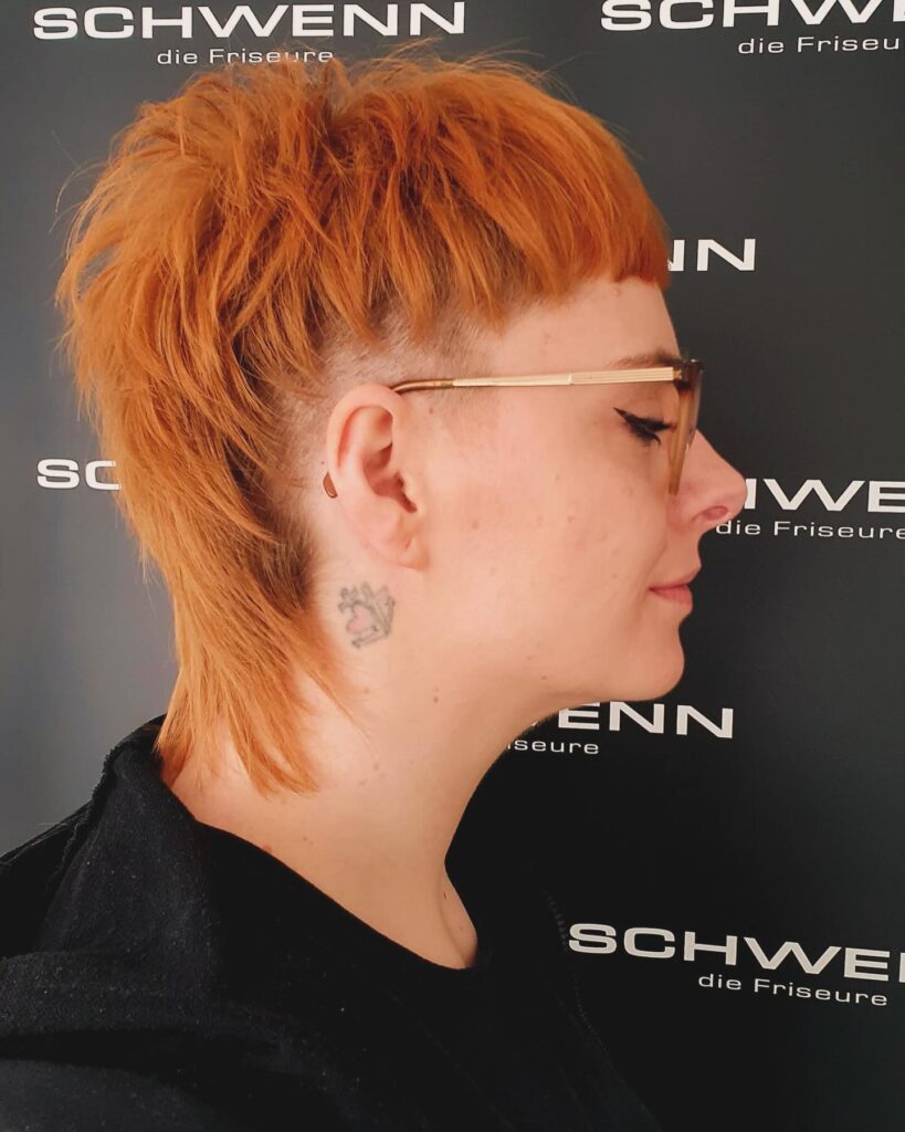 A side profile of a woman with a strawberry ginger wolf cut featuring a short fringe and shaved-temple undercuts.
