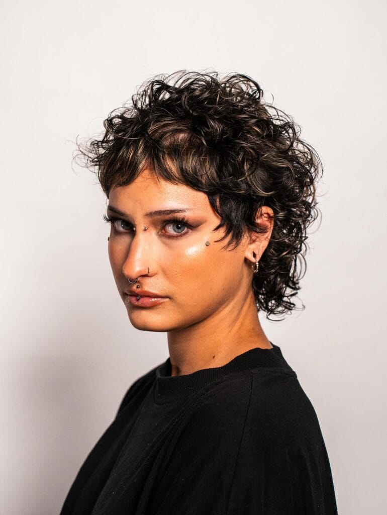 A woman with a dark brown curly pixie haircut featuring shaggy layers and subtle blonde highlights.