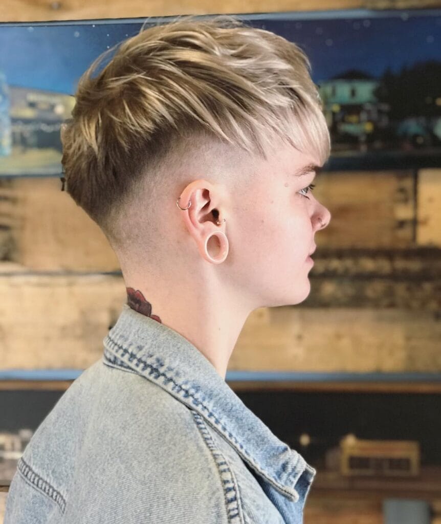 A person from a side profile showing a sandy blonde pixie haircut with a high skin fade and textured top layers.