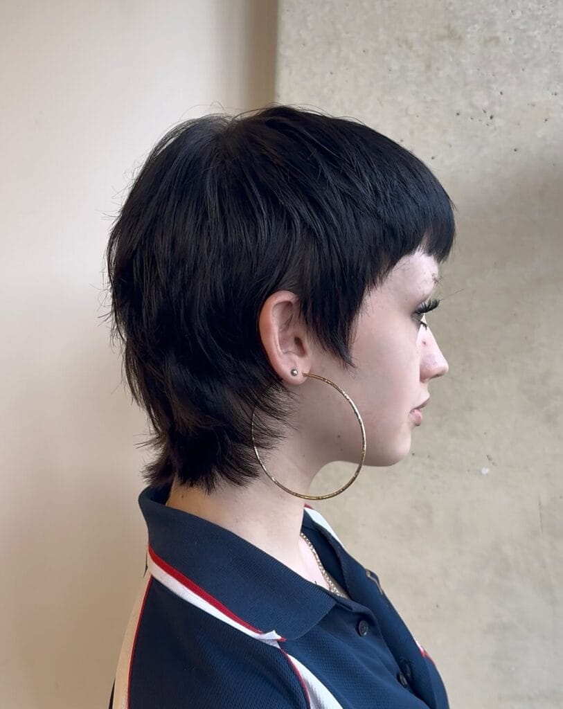 A side profile of a woman with a shaggy black pixie mullet featuring short blunt bangs and textured layers at the back.
