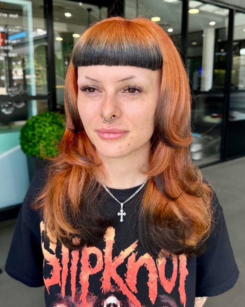 A woman with medium-length copper hair featuring blunt black micro bangs and dark-colored ends.