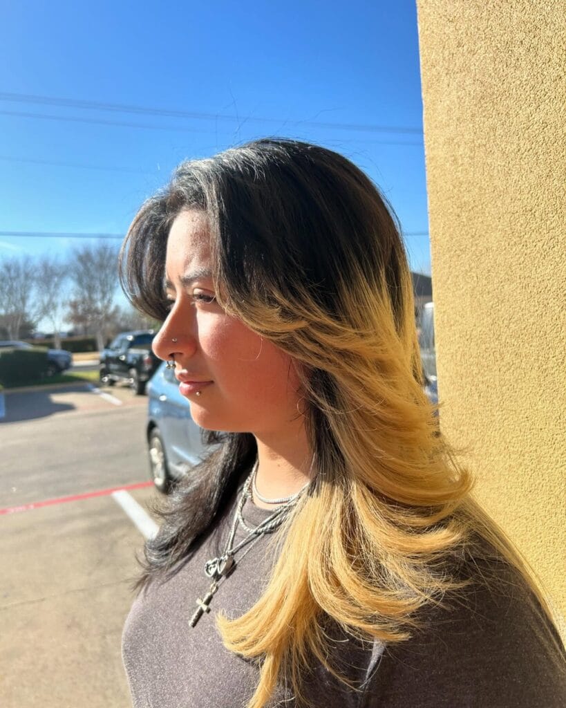 A woman with medium-length hair featuring a dark base and bright golden-blonde ombré with layered ends.