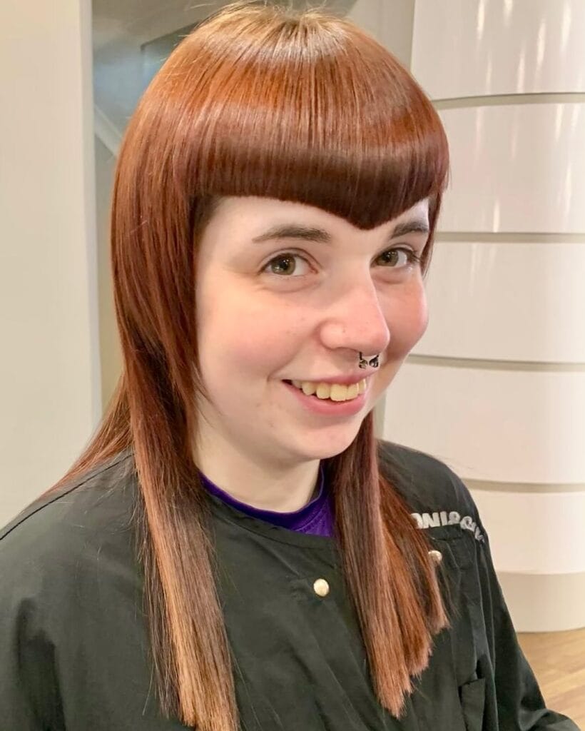 A woman with medium-length auburn hair featuring sharp V-shaped bangs and blunt side-framing pieces.