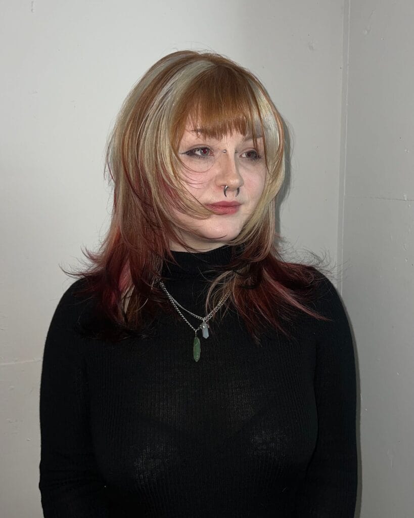 A woman with a medium-length layered shag featuring a mix of ginger and red tones with a bright blonde money piece.