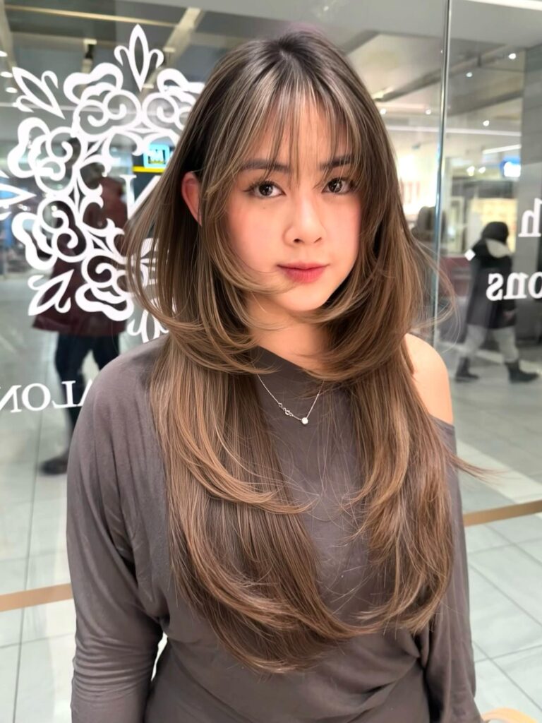 A woman with long, layered mushroom brown hair featuring caramel highlights and wispy bangs.