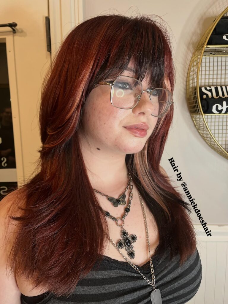A person with long auburn layered hair featuring a full fringe and subtle blonde peek-a-boo highlights.