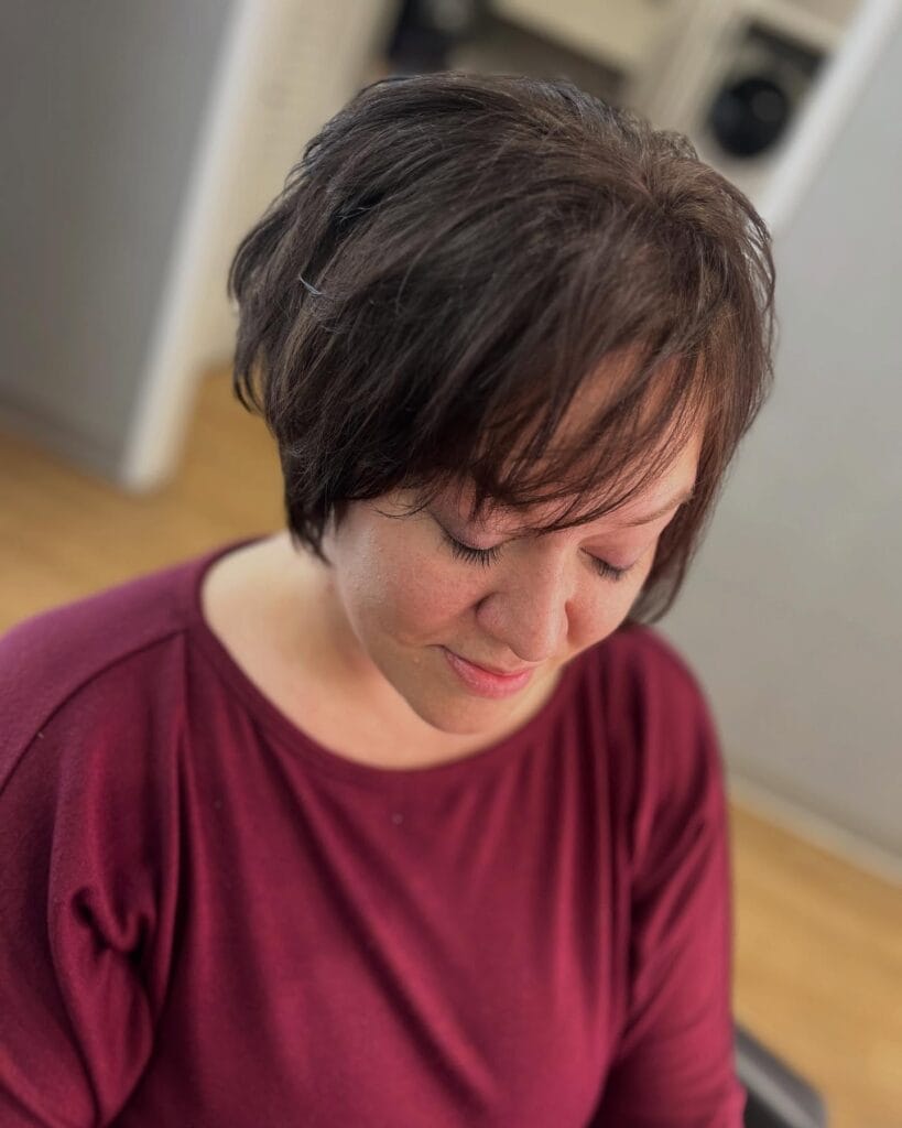 A woman looking down, showcasing a soft dark brown pixie cut with wispy bangs and gentle layers.