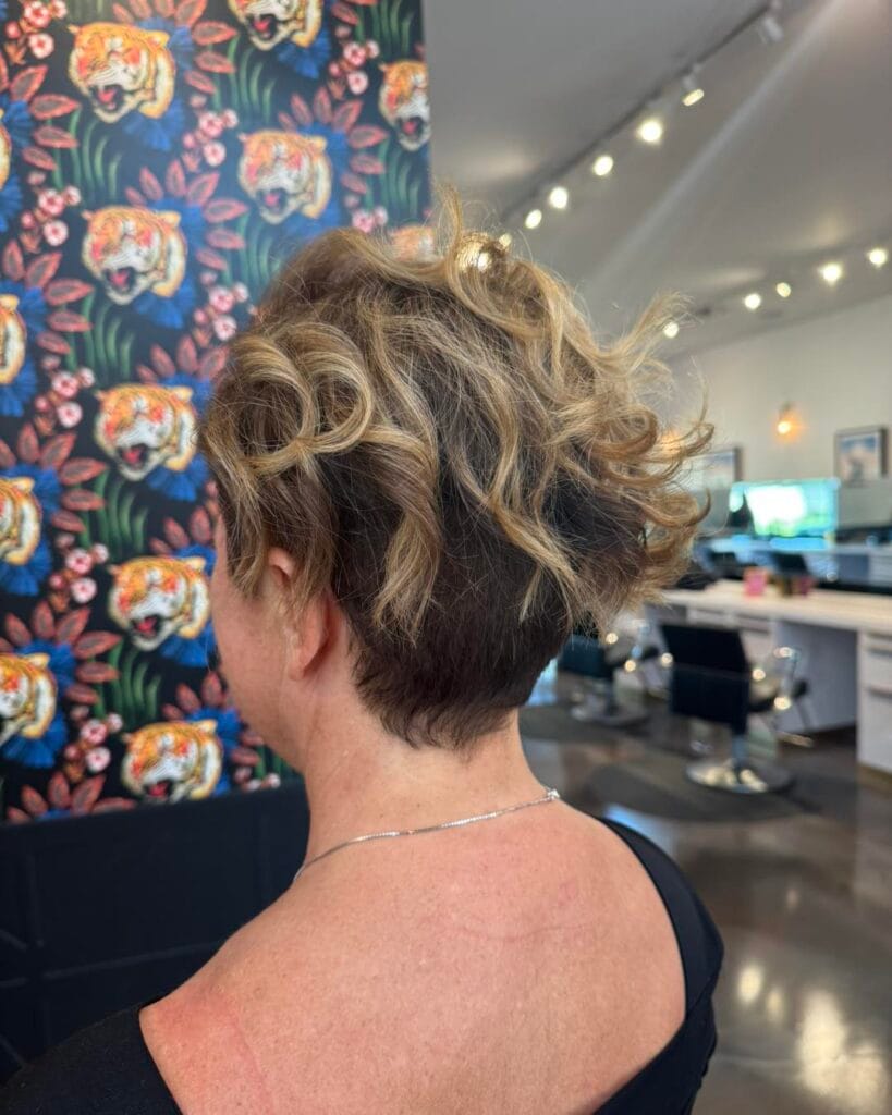 A back view of a woman with a short curly pixie cut featuring honey-blonde highlights and a tapered nape.