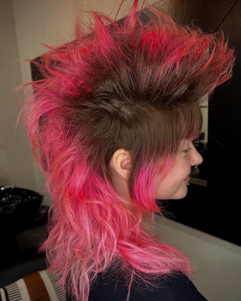 A woman with a voluminous hot pink mohawk mullet featuring tall textured spikes on top and long wavy pink hair in the back.