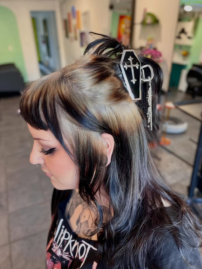 A woman with black hair and blonde skunk stripe panels styled half up with a decorative coffin hair clip.